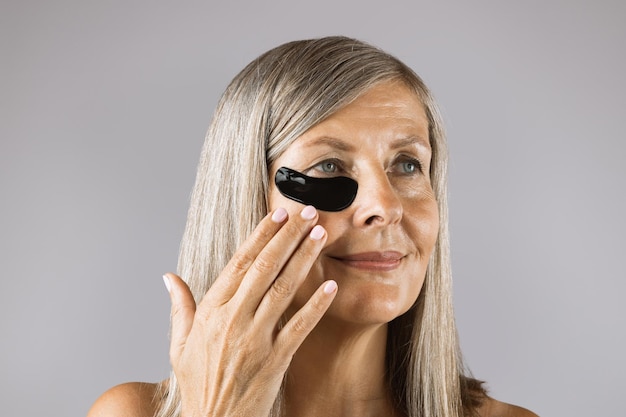 Aged woman applying black hydrogel patches under eyes