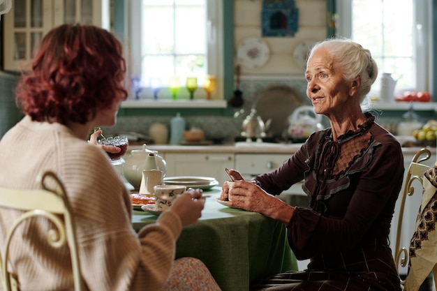 Aged woman in casualwear having tea and talking to her granddaughter