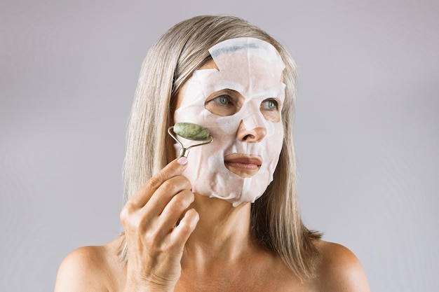 Foto donna anziana in maschera sul viso che fa massaggio con rullo di giada