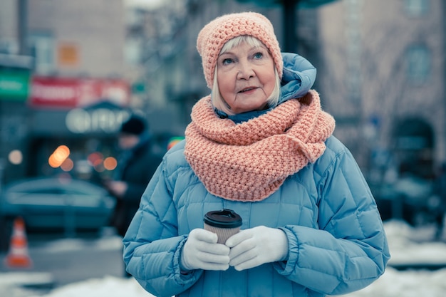 Фото Пожилая женщина в зимнем пальто, шляпе и шарфе, находясь на улице с картонной чашкой кофе