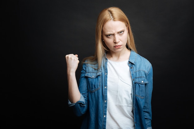 Aggressive decisive young woman expressing rage and showing fist while looking with anger and standing against black wall