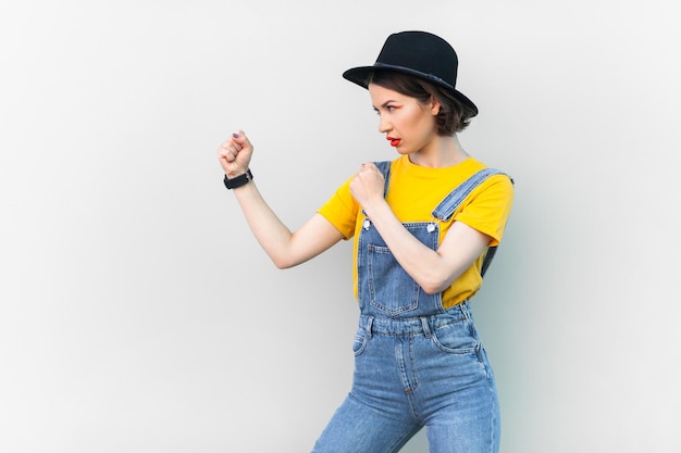 Aggressive hipster woman in overalls tshirt and hat keeps fists clenched being ready to fight