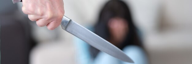 Aggressive man holds knife in hand against background of frightened woman. Husband threatens physical violence concept