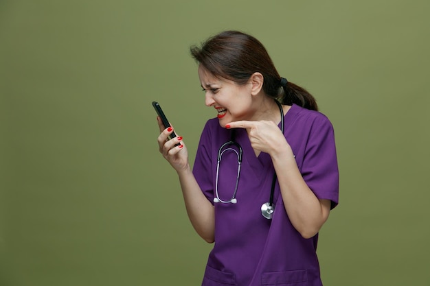 Aggressive middleaged female doctor wearing uniform and stethoscope around neck holding mobile phone looking at it pointing at it isolated on olive green background