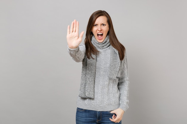 Aggressive woman in gray sweater scarf screaming showing stop gesture with palm isolated on grey background. Healthy fashion lifestyle people sincere emotions, cold season concept. Mock up copy space.