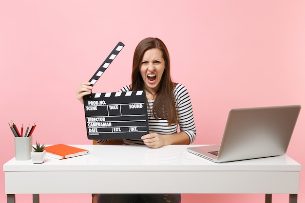 Aggressive woman screaming holding classic black film making clapperboard and working on project while sit at office with laptop 