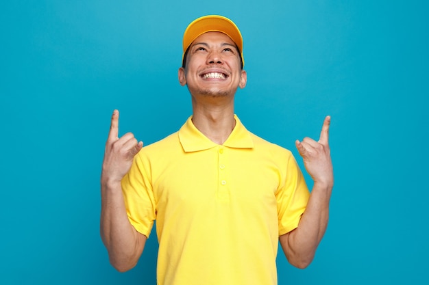 Aggressive young delivery man wearing uniform and cap looking up doing rock sign 