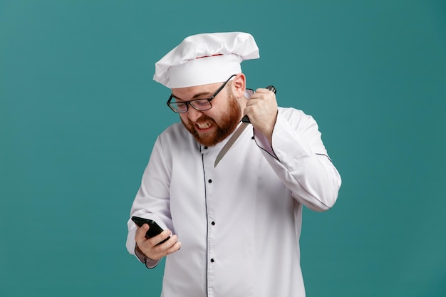 Aggressive young male chef wearing glasses uniform and cap holding and looking at mobile phone holding knife pointing knife at phone isolated on blue background