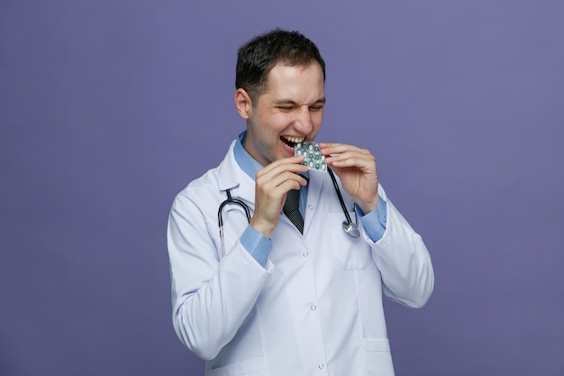 Aggressive young male doctor wearing medical robe and stethoscope around neck holding and biting pack of capsules with closed eyes isolated on purple background