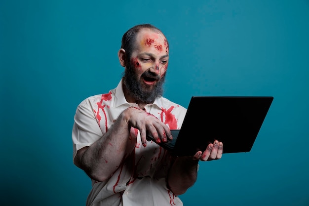 Aggressive zombie using laptop computer in studio, looking creepy and dangerous browsing internet while being spooky and apocalyptic. Brain eating corpse with bloody scars using social media.