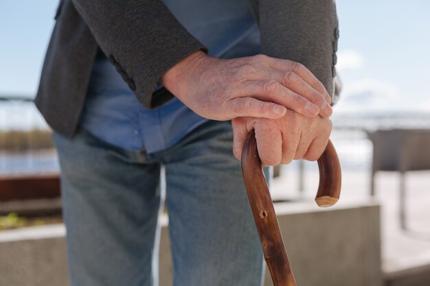 Aging concentrated old pensioner standing on the promenade and putting his hands on the stick and relaxing