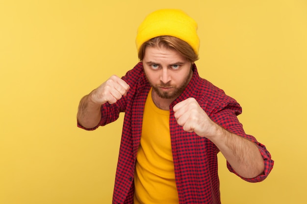Agressieve hipster bebaarde man in beanie hoed, geruit hemd met gebalde vuisten klaar om te boksen, ponsen met zelfverzekerde uitdrukking, vechtlust. studio-opname geïsoleerd op gele achtergrond