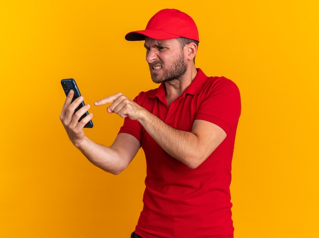 Agressieve jonge blanke bezorger in rood uniform en pet met kijkend naar en wijzend op mobiele telefoon