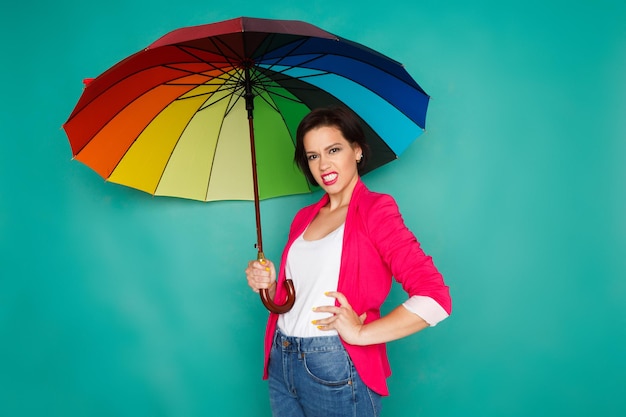 Agressive stylish girl in bright casual clothes under rainbow colored umbrella posing at azur studio background, copy space