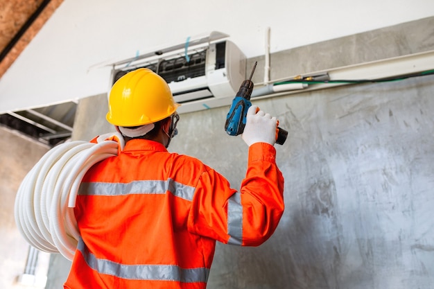 Air conditioner technician mechanic wearing mask and helmet to\
prevent disease, covid 19 currently using electric drill to install\
air conditioner.