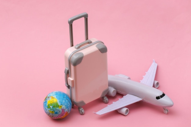 Air travel concept. Mini plastic travel suitcase, air plane, globe on pink background.