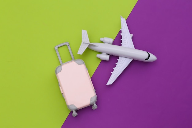 Air travel flat lay. Mini plastic travel suitcase and air plane on green purple background. Top view.