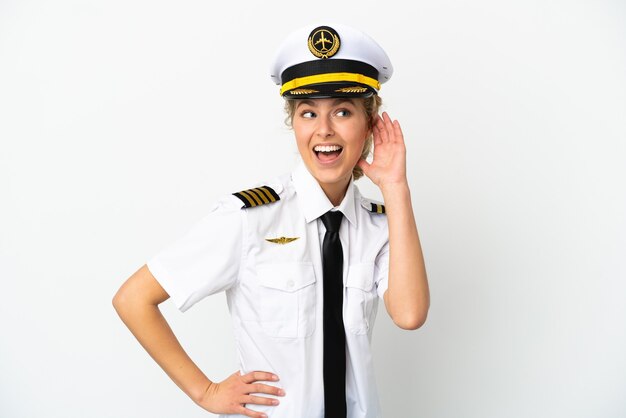 Airplane blonde woman pilot isolated on white background listening to something by putting hand on the ear