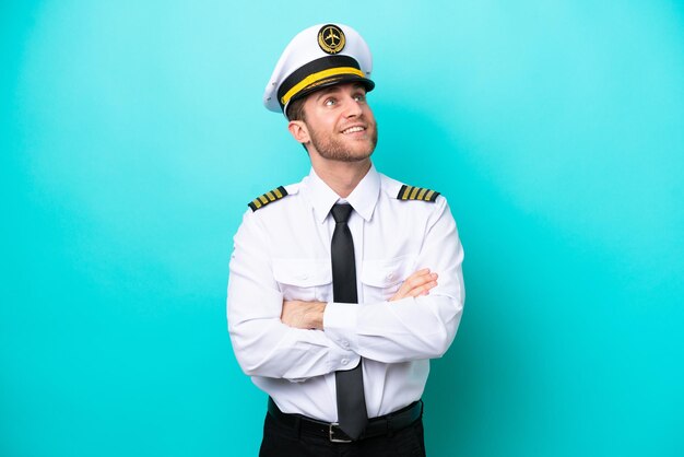 Airplane caucasian pilot isolated on blue background looking up while smiling