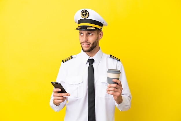 Pilota caucasico dell'aeroplano isolato su sfondo giallo che tiene il caffè da portare via e un cellulare mentre pensa a qualcosa