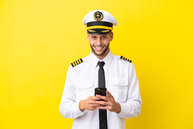 Pilota caucasico dell'aeroplano isolato su sfondo giallo che invia un messaggio con il cellulare