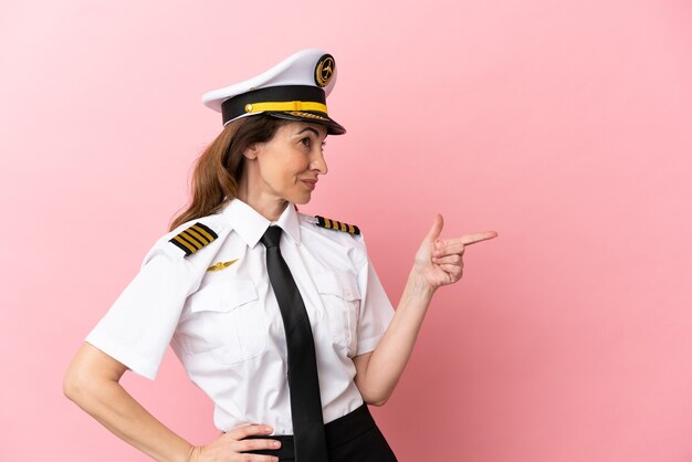 Airplane middle aged pilot woman isolated on pink background pointing finger to the side and presenting a product