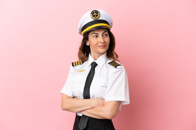 Женщина-пилот самолета средних лет изолирована на розовом фоне со скрещенными руками и счастлива