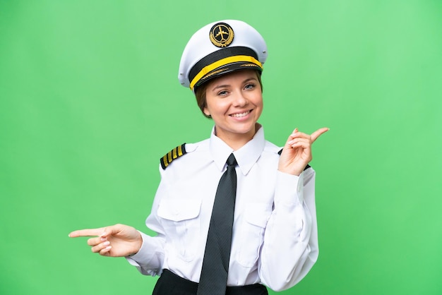 격리된 크로마 키 배경 위에 있는 비행기 조종사 여성이 측면과 행복을 손가락으로 가리키고 있습니다.