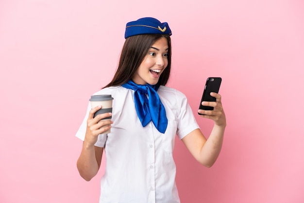 Airplane stewardess Brazilian woman isolated on pink background holding coffee to take away and a mobile