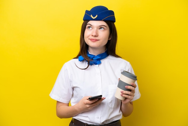 Airplane stewardess russian woman isolated on yellow background holding coffee to take away and a mobile while thinking something