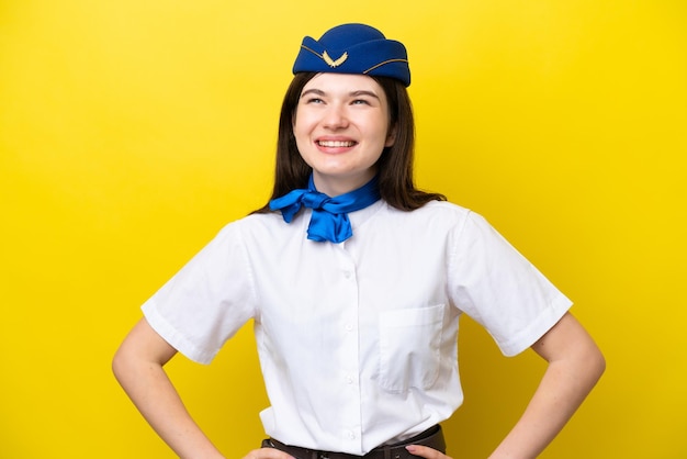 黄色の背景に分離された飛行機のスチュワーデスロシア人女性が腰に腕と笑顔でポーズをとる