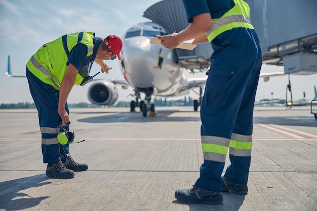 Foto lavoratore dell'aeroporto vestito in uniforme che indica l'aereo parcheggiato al suo collega maschio caucasico