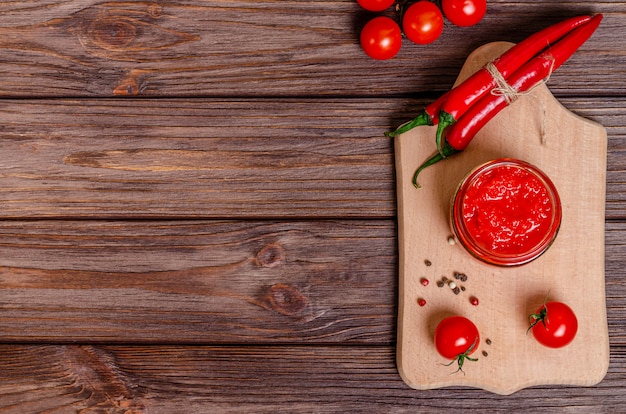 Ajvar or pindjur red vegetable spread or pepper paprika and tomato mousse in a glass jar on a cutting board on a rustic wooden table.