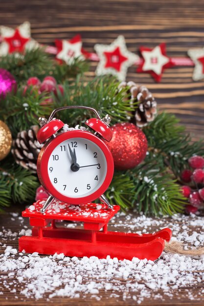 Alarm clock on little sledge with snow and Christmas tree on table on brown wooden background