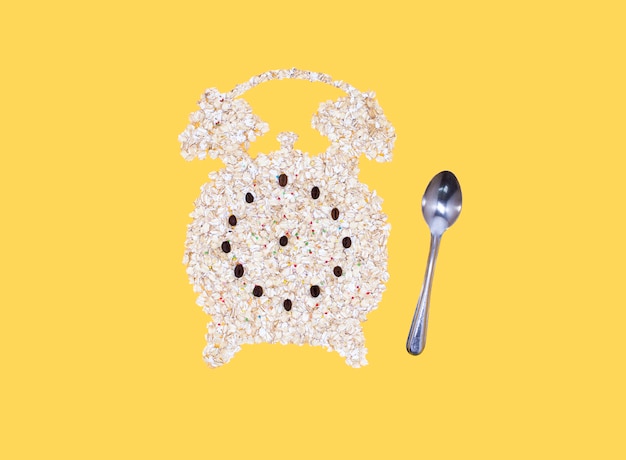Photo alarm clock of oatmeal and a metal spoon on a yellow background. healthy breakfast