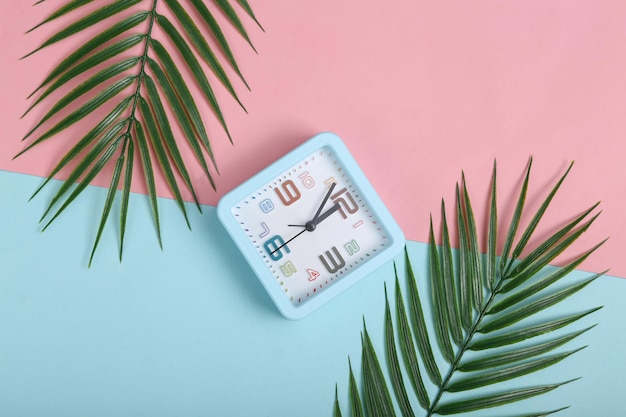 Alarm clock and palm leaves on pink blue background Top view Flat lay