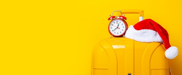 Alarm clock and Santa Claus hat lie on top of a yellow suitcase on a yellow background Banner