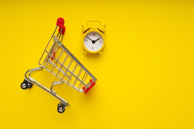 Alarm clock on the shopping cart on yellow background Shopping time concept