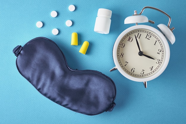 Alarm clock, sleeping mask, ear plugs and pills on blue background