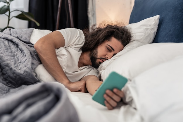 目覚まし時計。ベッドに横たわっている間、スマートフォンの画面を見ている眠そうなブルネットの男