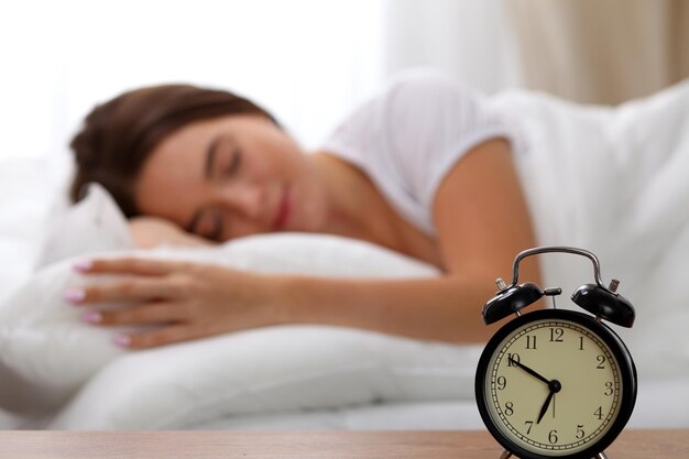 Alarm clock standing on bedside table has already rung early morning to wake up woman in bed sleeping in background. Early awakening, not getting enough sleep, oversleep, time line concept.