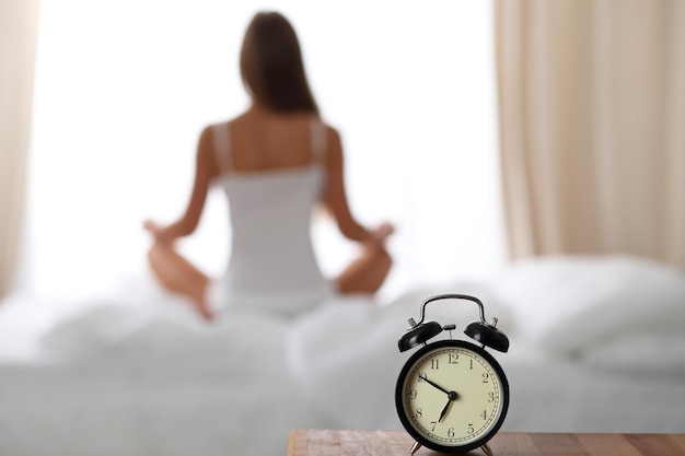 ベッドサイドテーブルに立っている目覚まし時計はすでに早朝に目覚めるために鳴っています女性はバックグラウンドでベッドでヨガをします早朝覚醒健康的なライフスタイル熟考の概念