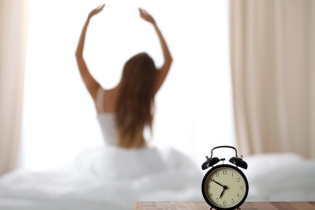 Фото Будильник, стоящий на прикроватной тумбочке, уже прозвонил рано утром, чтобы разбудить женщину, потягивающуюся в постели на заднем плане.