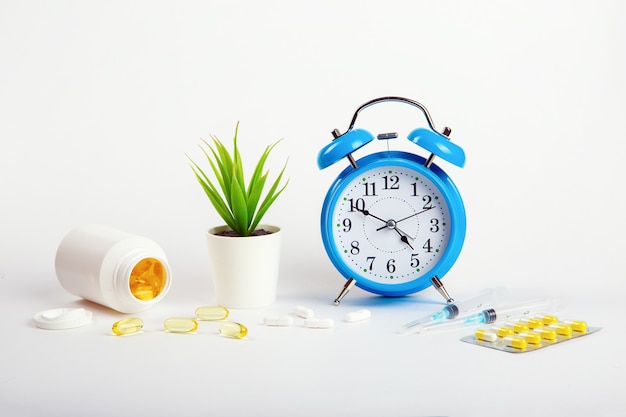 An alarm clock stands on a white wall shows the time of taking medications and next to it medical syringes and pills.