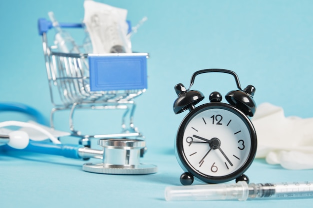 Alarm clock syringe and ampoules and shopping trolley on blue background stethoscope face mask health care and buying medicine concept