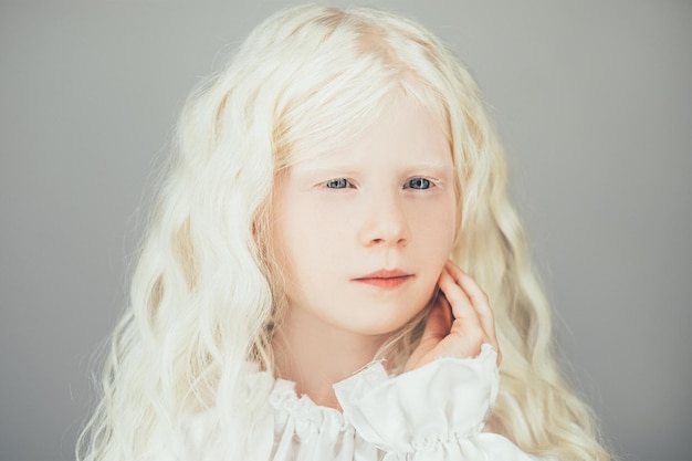 Albino beauty. Sweet child portrait. Pure innocence. Adorable thoughtful blonde small girl with white eyebrows curly hair blue eyes in vintage blouse isolated on gray background.