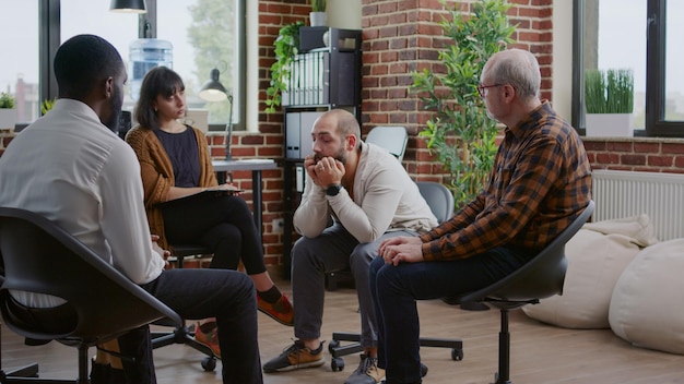 Alcohol addict sharing anger management issues with people at aa rehab meeting. Nervous man with mental health problems talking to patients and psychologist in circle at group therapy.