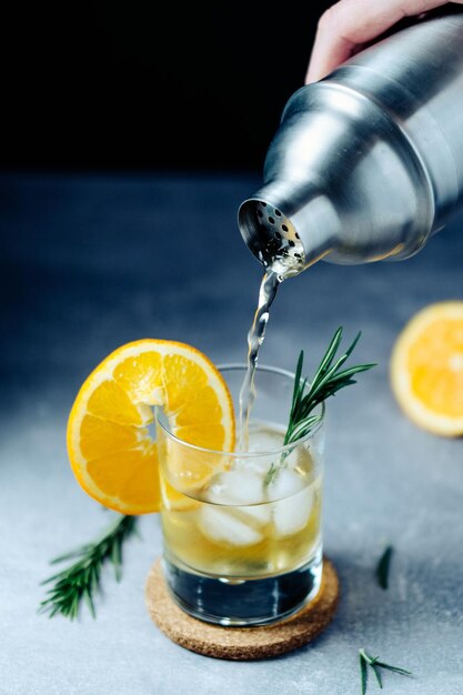 Alcohol cocktail pouring from shaker into glass with orange slice rosemary and ice on wooden desk