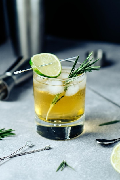 Alcohol cocktail with lime slice rosemary ice and cocktail shaker on black background Party time