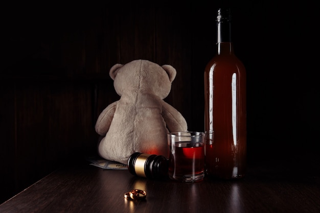 Alcohol en echtscheiding concept. Teddybeer, ringen en fles met glas in een donkere kamer.